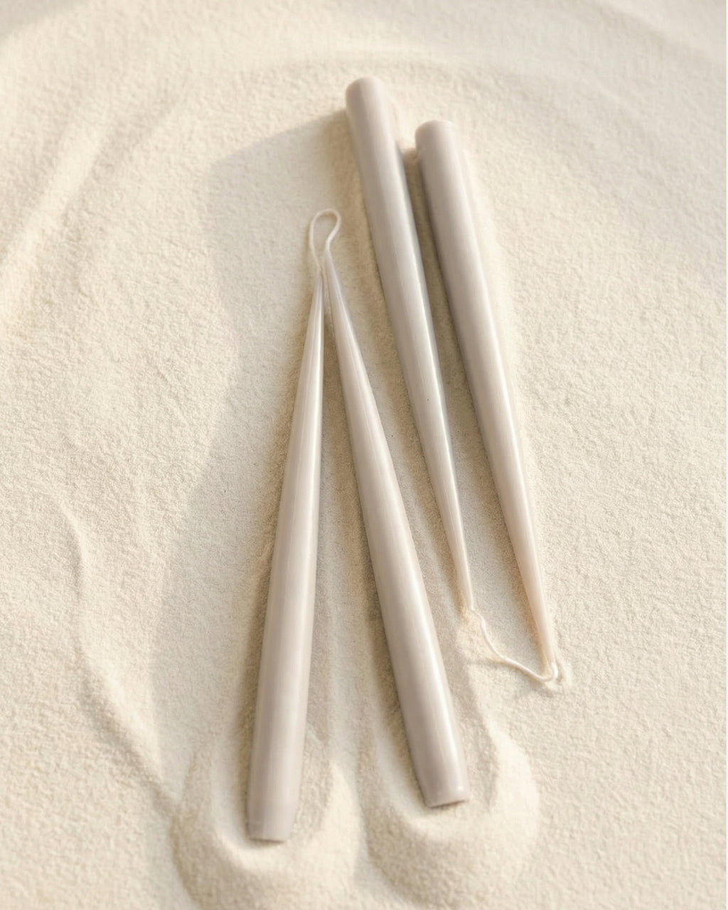 Hand-finished taper candles in grey shade arranged on a textured neutral surface (sand)