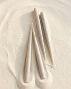 Hand-finished taper candles in grey shade arranged on a textured neutral surface (sand)