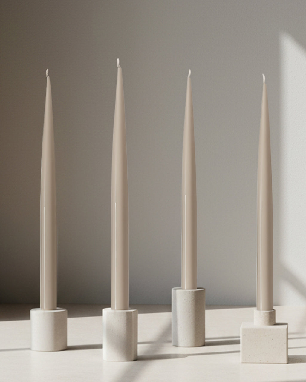Four PLAMYA taper candles in Sand Ash (grey) shade placed in stone candle holders on a neutral surface