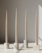 Four PLAMYA taper candles in Sand Ash (grey) shade placed in stone candle holders on a neutral surface