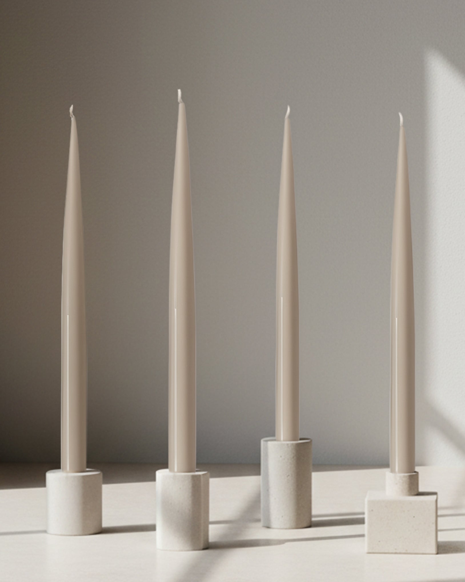 Four PLAMYA taper candles in Sand Ash (grey) shade placed in stone candle holders on a neutral surface