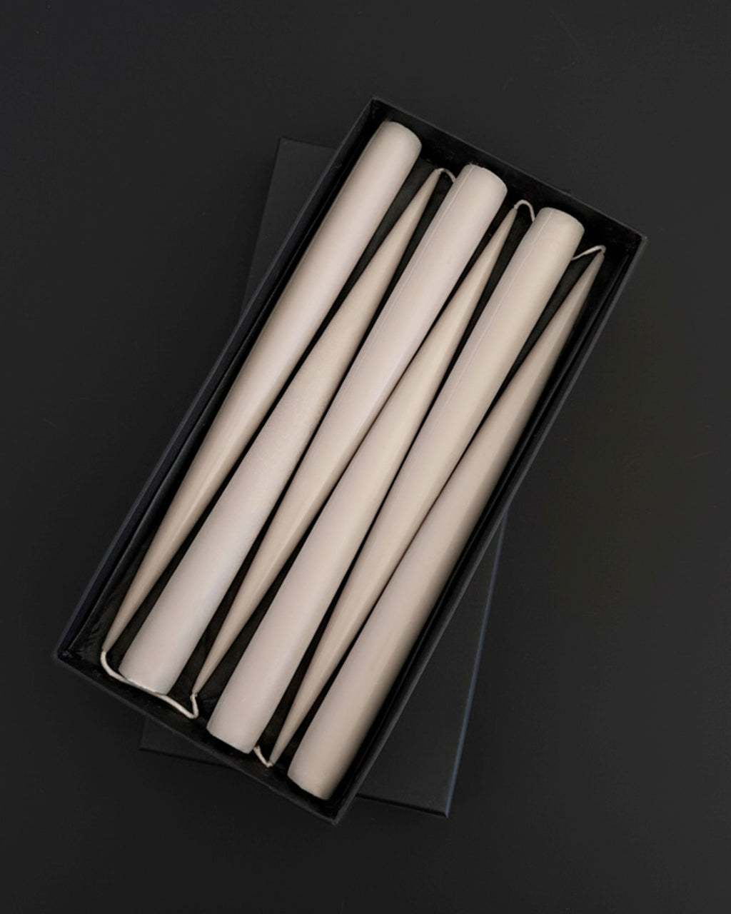 Set of six PLAMYA taper candles in Sand Ash (grey) shade arranged in a black box on a dark background