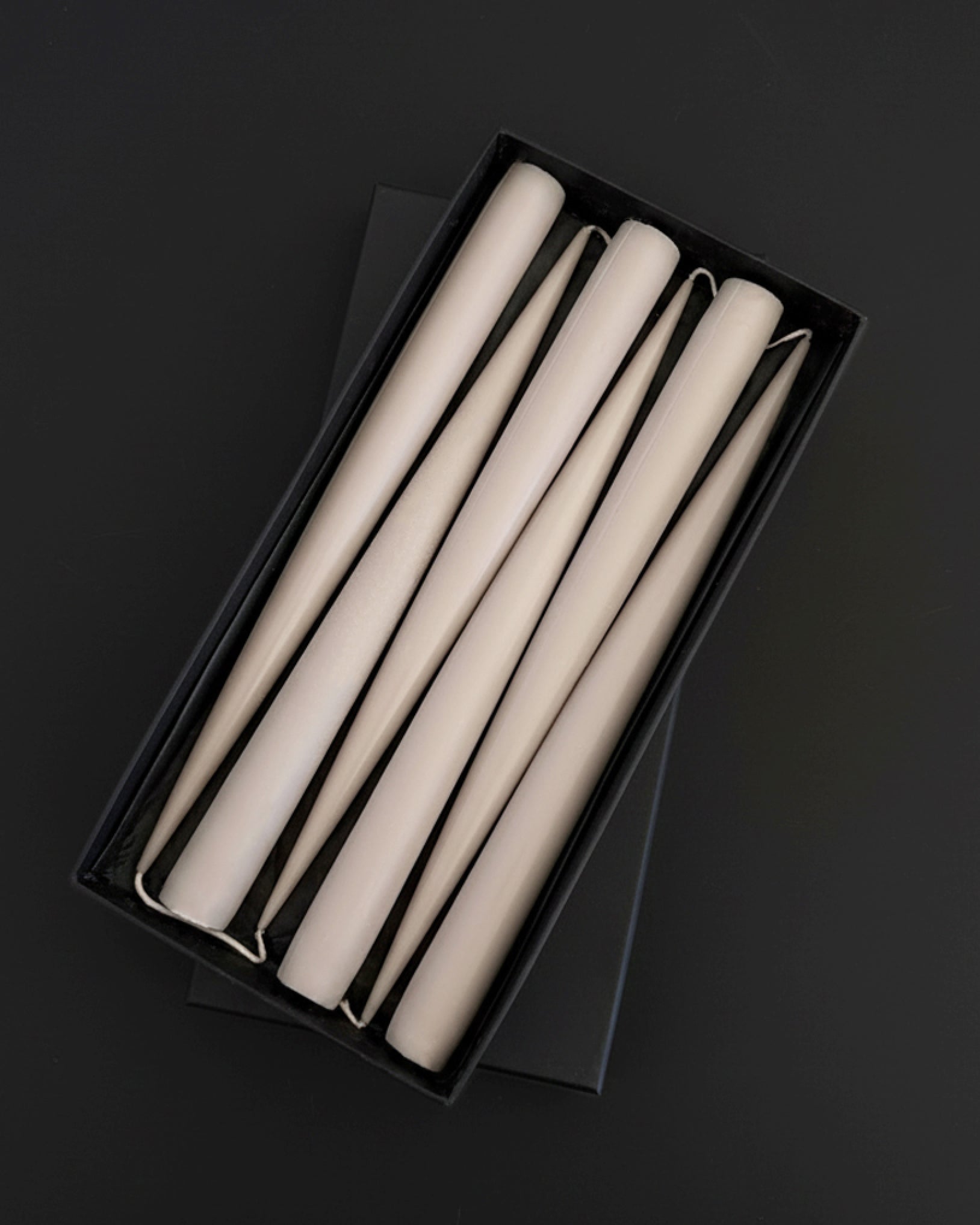 Set of six PLAMYA taper candles in Sand Ash (grey) shade arranged in a black box on a dark background