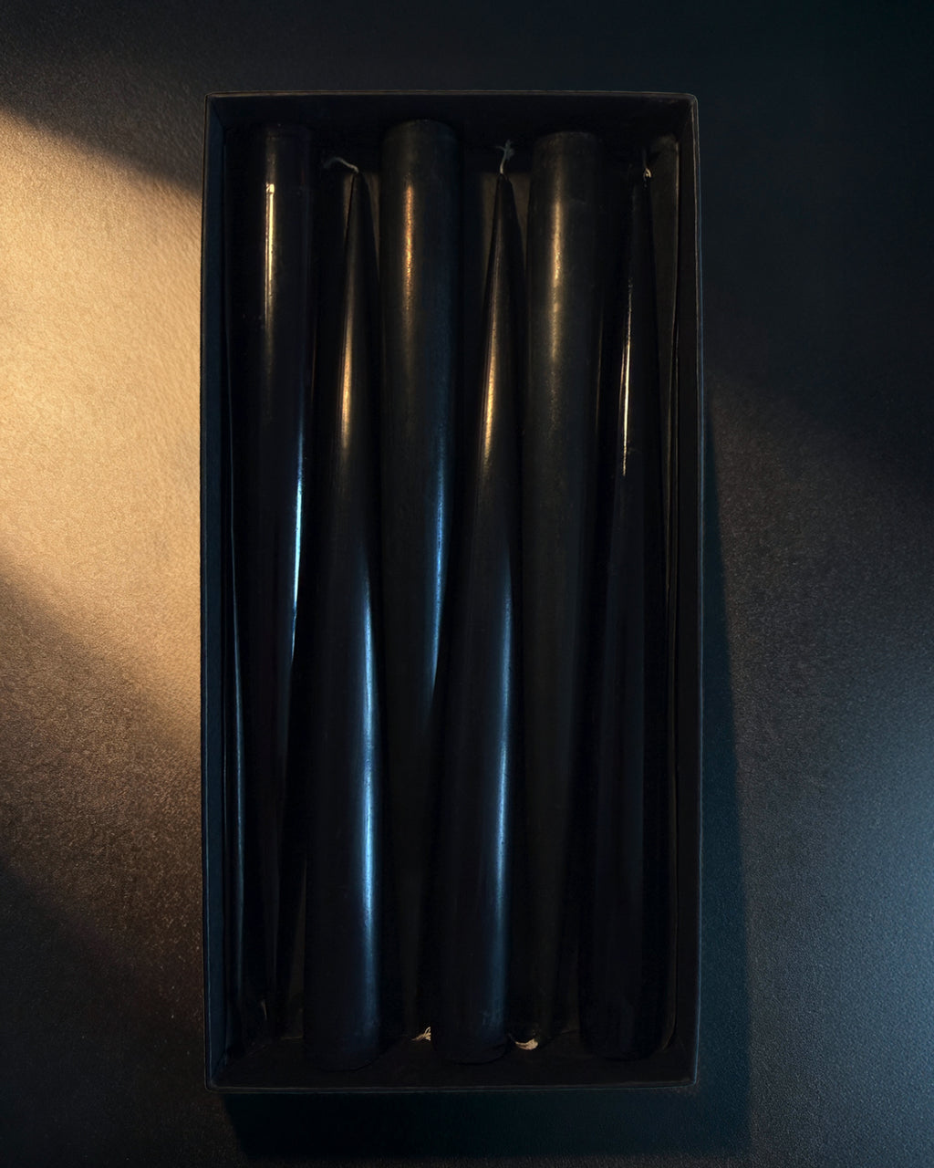 Set of black PLAMYA taper candles arranged in a black box on a dark background